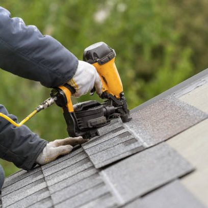 Unrecognizable roofer worker in uniform work wear using air or pneumatic nail gun and installing asphalt or bitumen tile on top of the roof under construction house